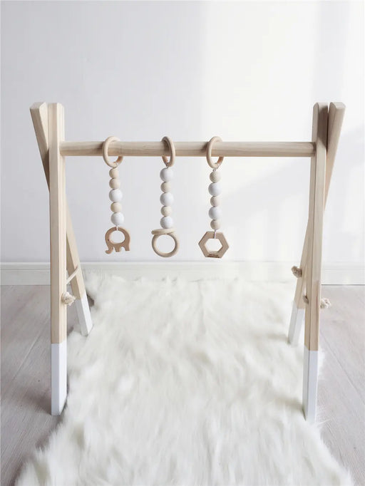 Ett minimalistiskt Babygym från Bebisgrossisten.se med vita accenter sitter på en fluffig vit matta. Perfekt för tidig motorisk utveckling, detta babygym i trä har tre hängande leksaker: en träelefant, en ring med vita pärlor och en hexagonform för att engagera och stimulera ditt barns sinnen.