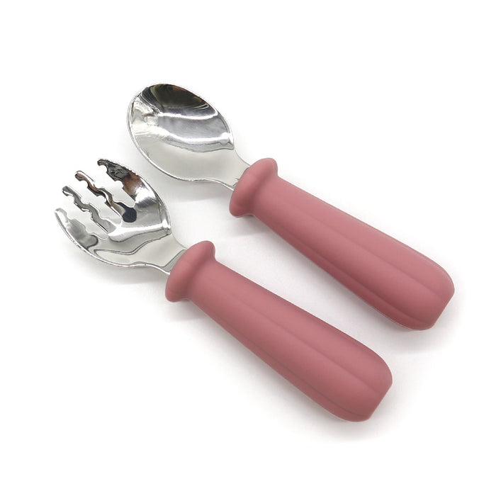 Två redskap från Toddlers Of Swedens Barnbestick Med Silikonskaft visas: en sked och en spork. Båda har glänsande metallhuvuden och rosa, texturerade silikonskaft-handtag designade för enkelt grepp. Spork har tre pinnar, och båda verktygen verkar väl lämpade för småbarn som lär sig att självmata med BPA-fria material.