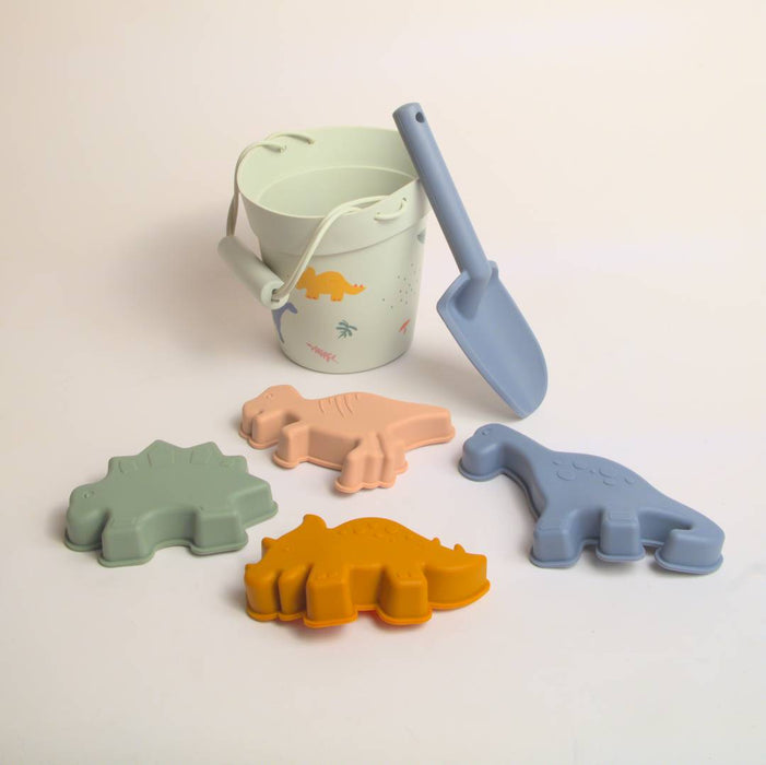 Silikon Strandleksaker-setet av Toddlers Of Sweden har en vit hink med ett handtag och dinosaurieillustrationer, en blå plastskyffel och fyra frukt före detta dinosaurieformade sandformar tillgängliga i grönt, rosa, orange och blått, allt tillverkat av återvunnet silikon. Hela setet visas vackert på en vit bakgrund.