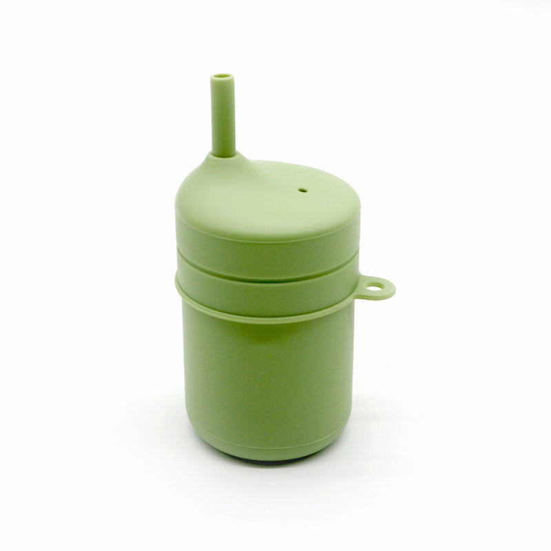 En TOS Silikon Pipmugg Sugrör från Toddlers Of Sweden, med en grön cylindrisk design med ett monterat lock som inkluderar ett inbyggt sugrör och ett litet handtag på sidan, allt mot en vit bakgrund. Den eleganta och minimalistiska pipmuggen är tillverkad av BPA-fritt material som ser ut att vara plast eller silikon.